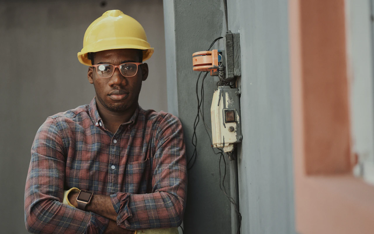 An electrician in a yellow hard hat and plaid shirt standing with folded arms beside an exterior meter box.