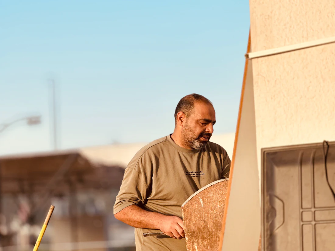 A worker in a tan t-shirt carrying a sheet of board past the side of a building.