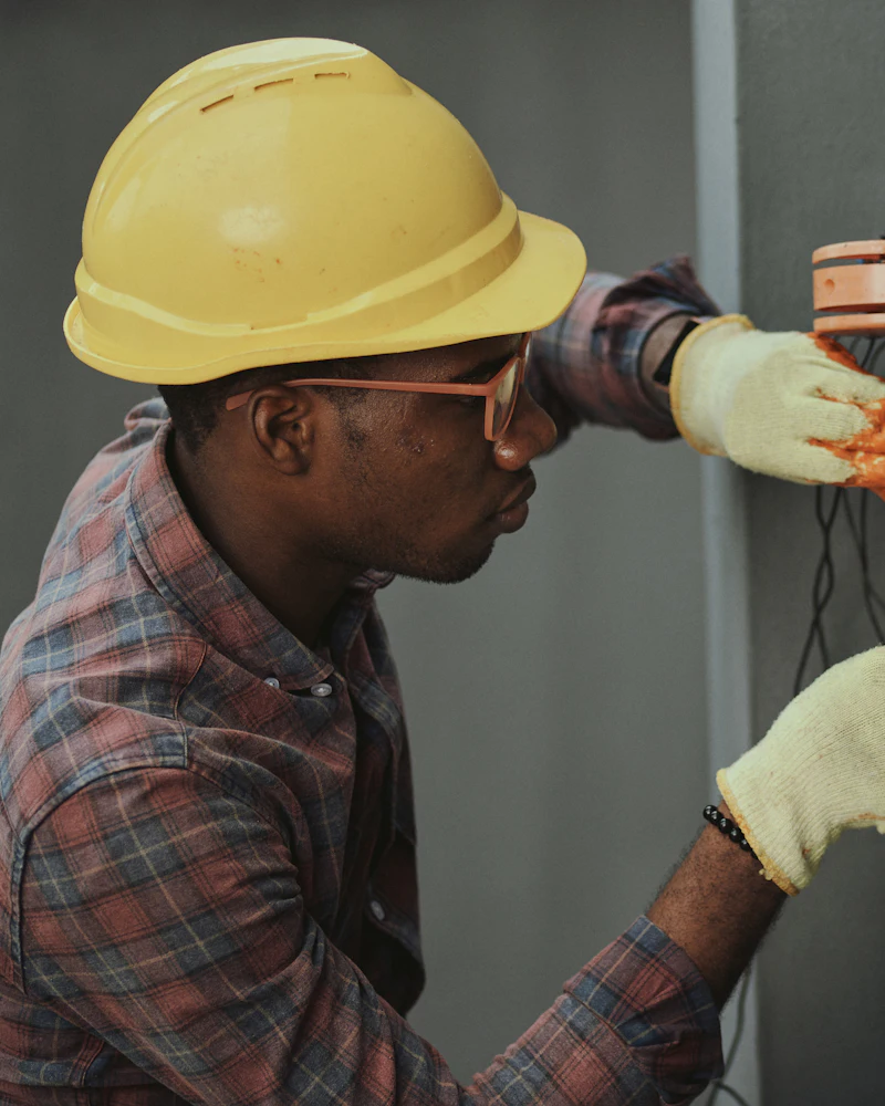 An electrician in a yellow hard hat and work gloves landing wires in a box on a wall.