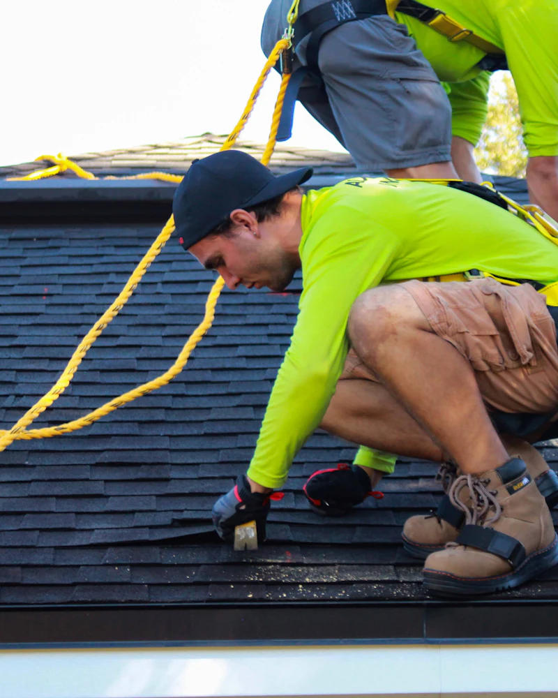 A roofer in a high-visibility shirt kneeling on a shingled roof with a nail gun, a yellow rope running across the slope to a second worker above.